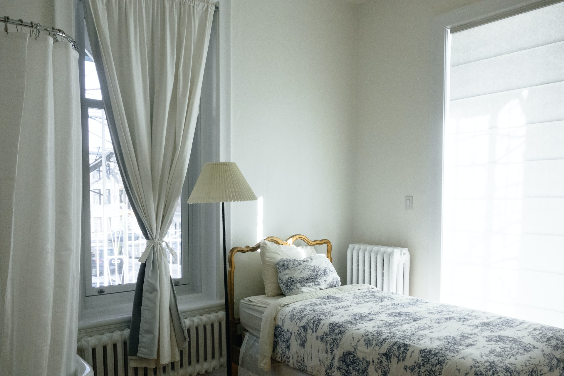 A small room with a bed with printed bedsheet, a white floor lamp and a grilled window
